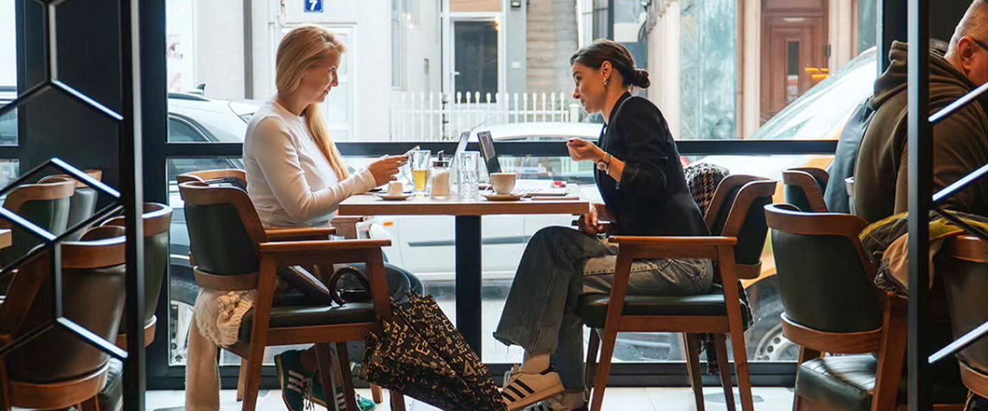 Two women working at a table by a large café window, with laptops and coffee, the street outside