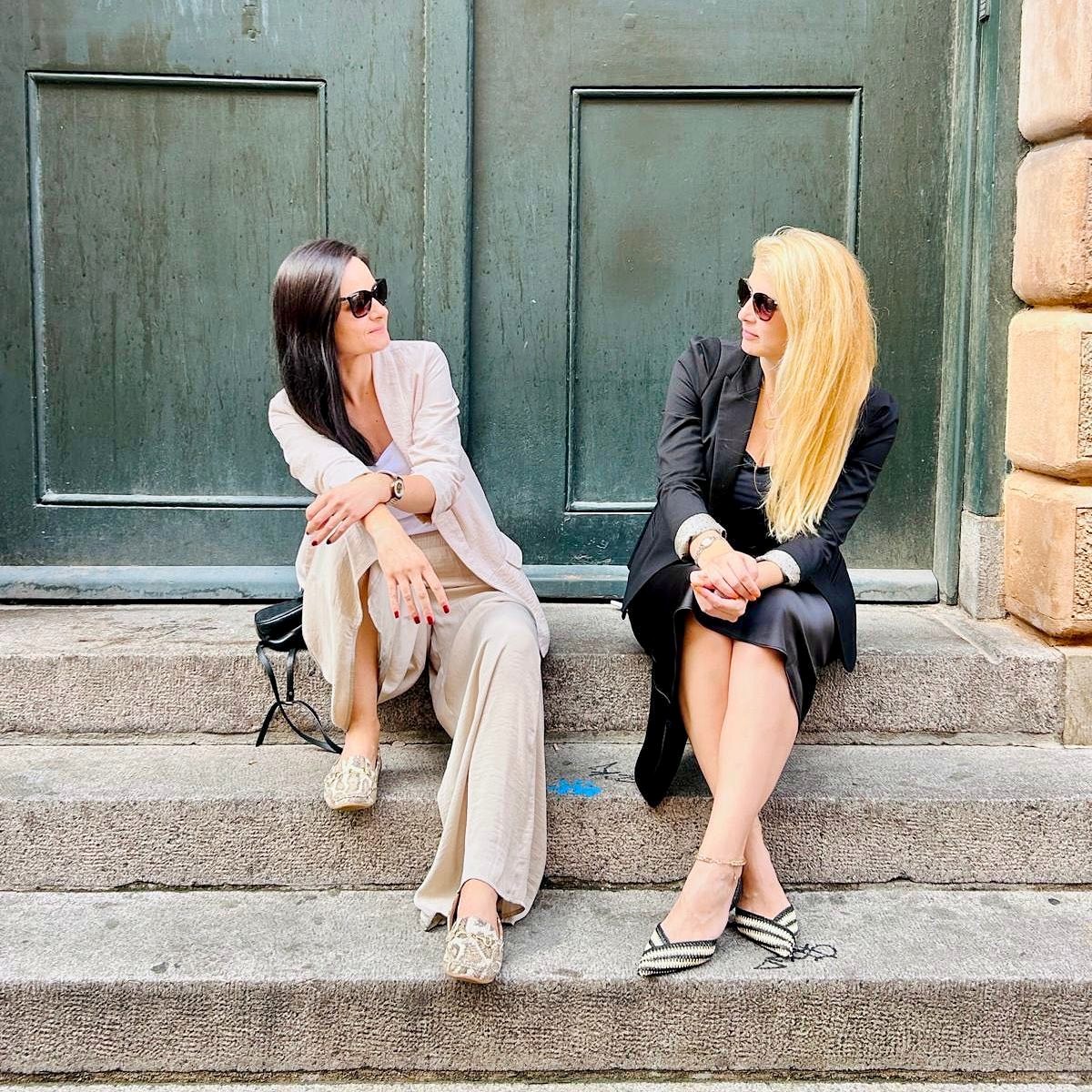 Maja and Marija in sunglasses, sitting on stone steps in front of large green doors and looking at each other