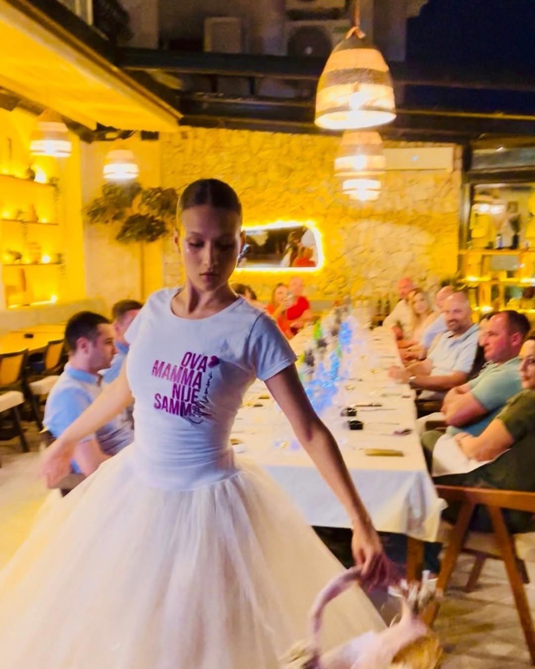 A performer in a white tutu dances between long banquet tables while guests watch, under warm restaurant light